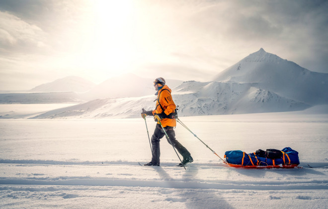 Participant pulling a sled through snow and snow-covered mountains in background