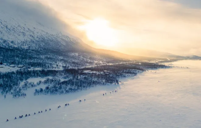 Aerial view of cyclists riding across a frozen snow-covered landscape at sunrise, surrounded by mountains and soft winter light.