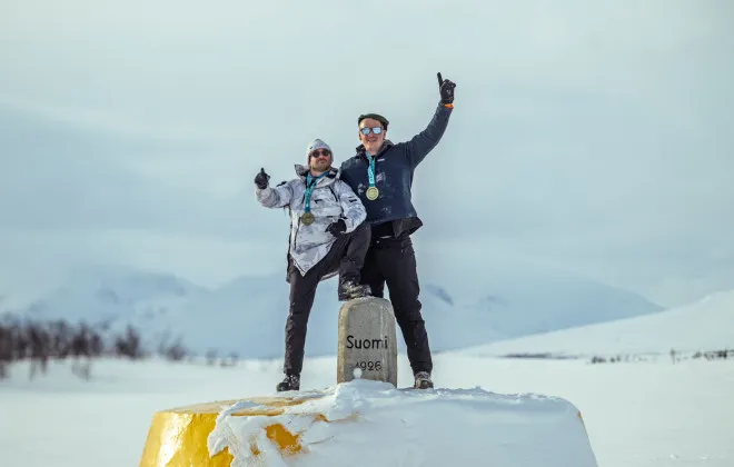 Two participants wearing medals standing on a snow-covered yellow marker in a winter mountain landscape, celebrating an achievement in snowy conditions.