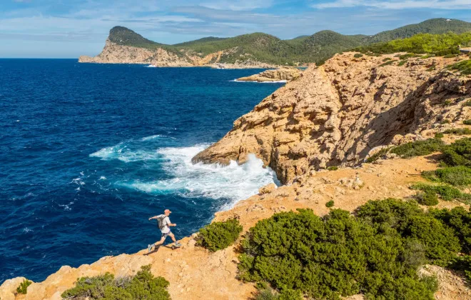 An event participant runs along the edge of a cliff beside a deep blue sea, with orange-red rock, patches of greenery, and a sunny sky above