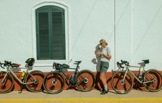 A blonde woman stands in front of a white wall, drinking from a mug, with three bicycles leaning against the wall behind her