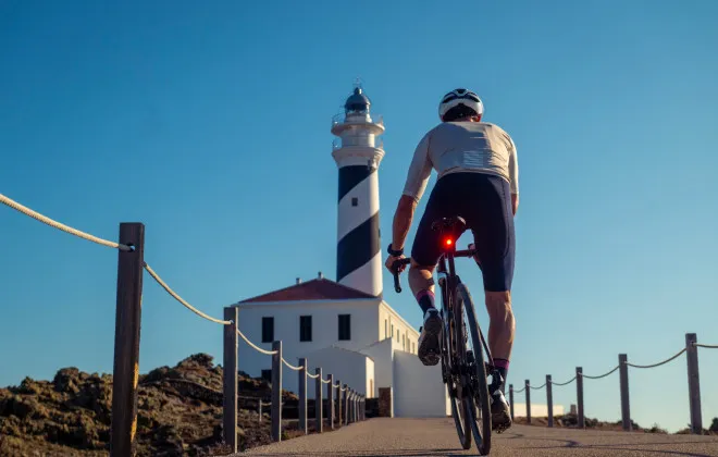 A cyclist riding along a path towards a lighthouse and a clear blue sky above