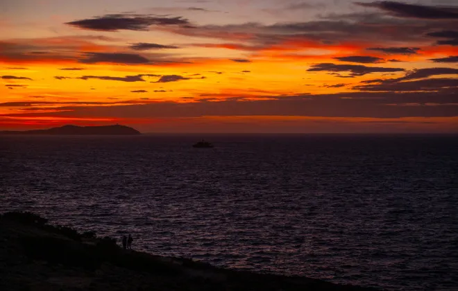 Two silhouettes look out onto a dark sea, with an orange and red sky above