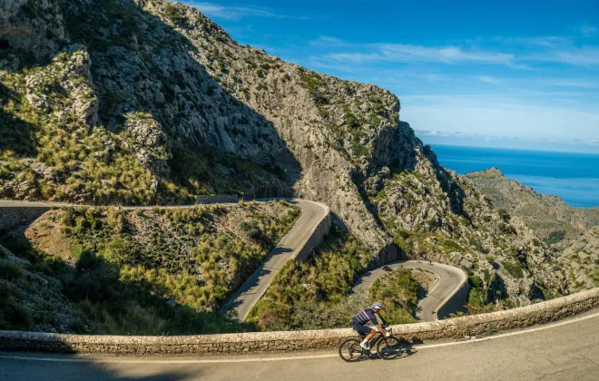 A lone cyclist rides along a winding road, with mountainous terrain, the sea, and a bright blue sky visible in the distance