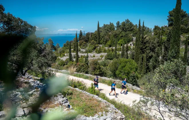 Three participants run along a dusty road, with greenery either side and the bright blue sky above