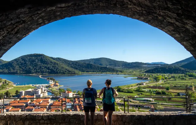 Two women stand under an arch, looking out onto buildings, water and greenery
