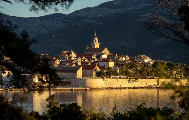 Buildings enclosed by a stone wall sit beneath mountainous scenery, with water stretching around