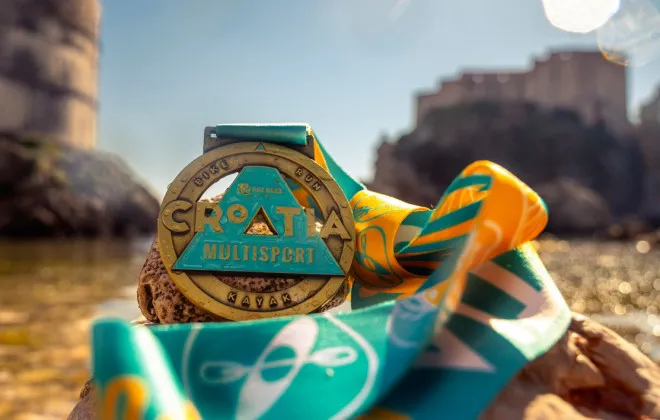 A Croatia Multisport medal with a colourful ribbon rests on rocks beside the water, with sunlit buildings blurred in the background