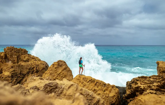 Woman standing on a sandy rock, with the sea splashing against it