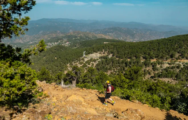 Man running along sandy and rocky terrain with greenery and mountains in the distance