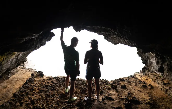 Two participants stand inside a rocky cave entrance, silhouetted against the bright daylight outside