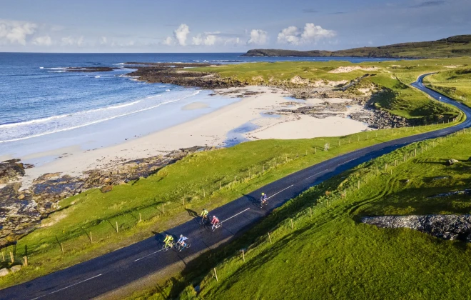 Cyclists riding along coastal road, with sandy beach to one side and green field to the other