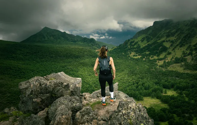 Woman stood on rock, looking out to forest and mountains with a cloudy sky above