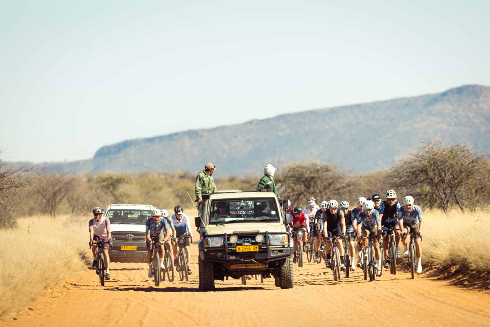 Cyclists and two vehicles moving along dusty road beneath sun